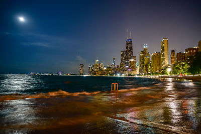 Illuminated buildings by sea against sky at night