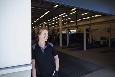 Smiling female mechanic looking away while leaning on wall at repair shop