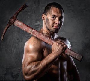 Portrait of shirtless muscular worker holding pick axe against wall