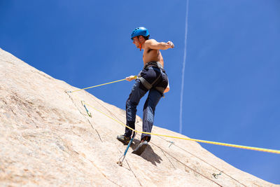 Low angle view of young man climbing rock against blue sky during sunny day