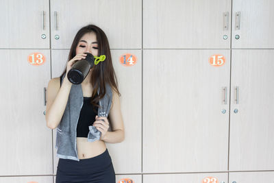 Woman drinking water from bottle while standing against locker