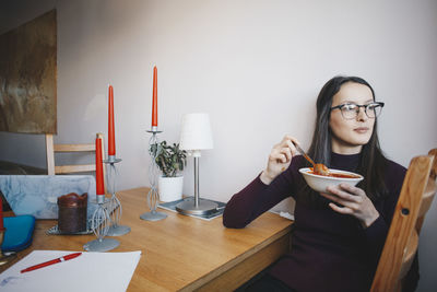 Young woman eating tomato soup sitting on chair by table against wall in dorm