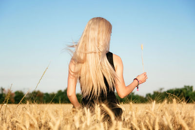 Rear view of woman holding plant while standing in wheat farm against clear sky