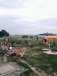 People relaxing on chairs at grassy field against sky