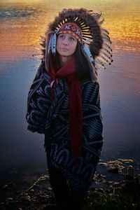 Thoughtful woman wearing feather headdress while looking away by lake during sunset