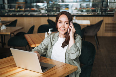 Adult business woman with long hair in stylish shirt working on laptop using mobile phone in cafe