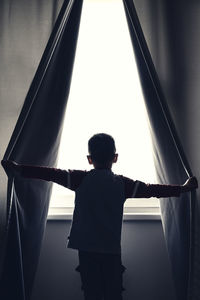 Rear view of boy holding curtains while looking through window at home