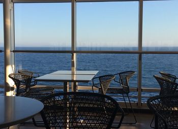 Empty chairs and tables by sea against sky seen through glass window