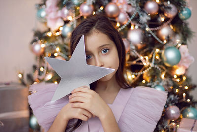 Beautiful baby girl with big eyes in a pink dress is sitting at the christmas tree at home hold star