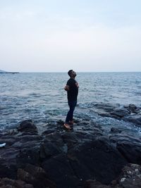 Side view of young man standing on rocky seashore against sky during sunset