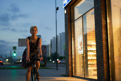 Woman in dress rides bicycle in night city near store