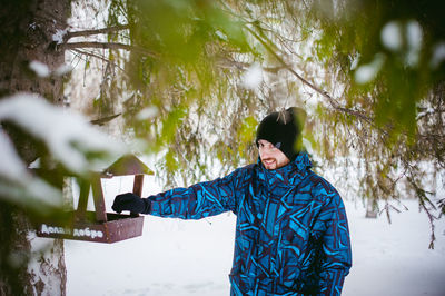 Portrait of young man standing by bird feeder on tree during winter