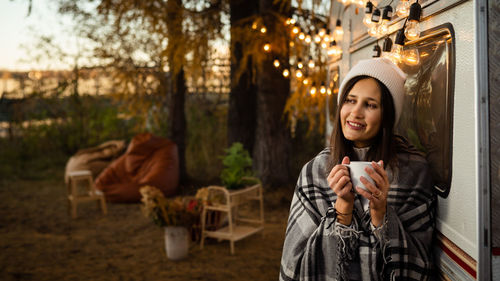 Caucasian woman in a knitted hat wrapped in a plaid and drinks a warming drink outdoors. 