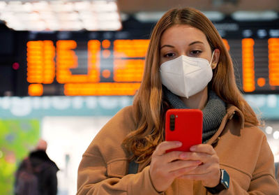 Woman wearing face mask using smartphone sanitary passport during pandemic and timetables boards