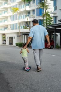 Father walking with girl riding push scooter on road in city