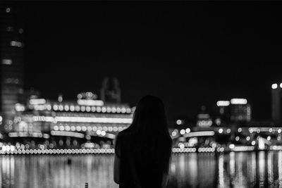 Rear view of woman standing by lake against illuminated city at night