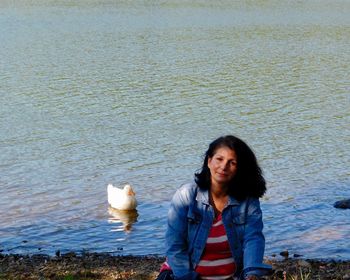 Portrait of woman sitting at lakeshore while duck swimming in lake