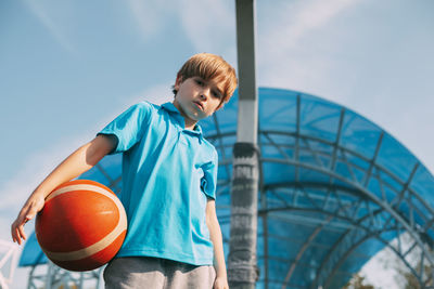 Low angle portrait of boy with basketball standing against sky