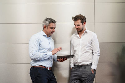 Smiling male entrepreneurs with digital tablet discussing about plan while leaning on wall