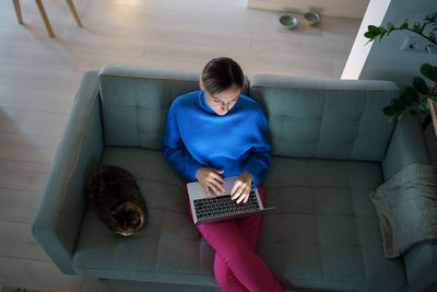 Top view caucasian woman with laptop on lap typing post for own blog sits on couch near with cat
