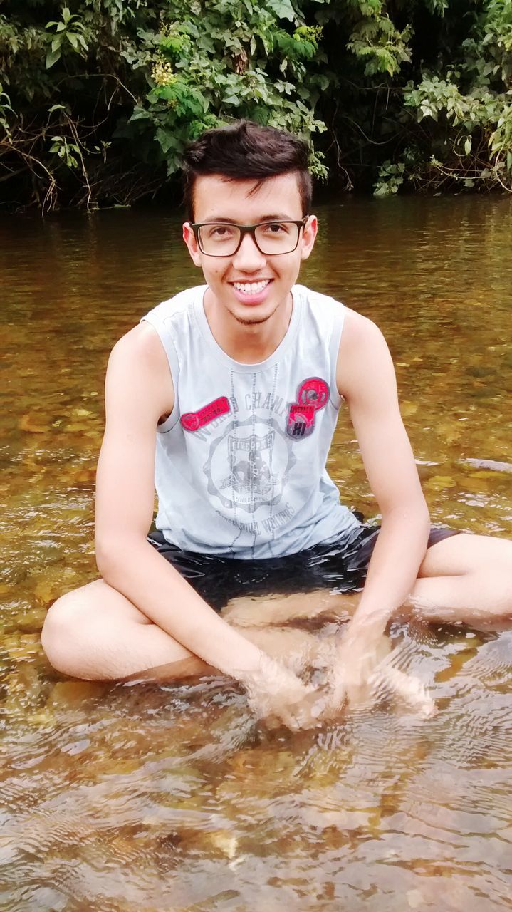 water, person, lifestyles, leisure activity, looking at camera, portrait, sitting, front view, smiling, holding, refreshment, tree, young adult, relaxation, lake, childhood, day, outdoors