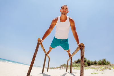 Low angle view of man exercising on parallel bars at beach against blue sky