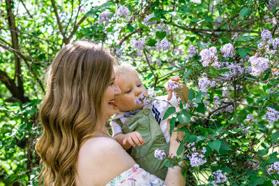 Portrait of young caucasian mother who is holding her son and touching flowers of lilac tree. 