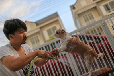 Low angel view of man bathing dog against fence