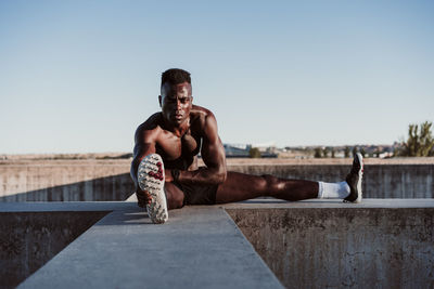 Sportsman doing stretching exercise while sitting on wall against clear sky during sunny day