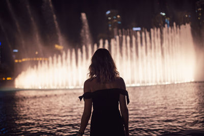 Rear view of woman standing against illuminated fountain in city