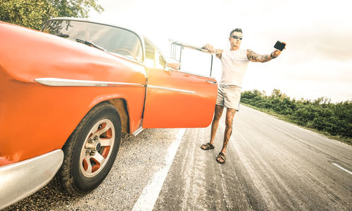 Man taking selfie while standing by red vintage car on road