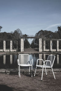 Empty chairs by lake against sky