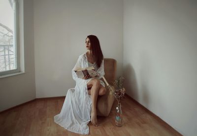 Young fashion model sitting with book on chair against wall