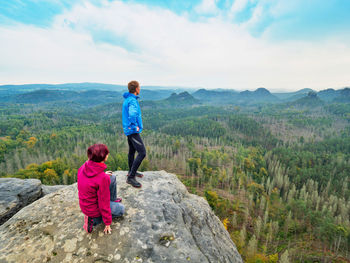 Boy and girl tourists stay on cliff and think. forest in valley with dry spruce trees. dreamy land