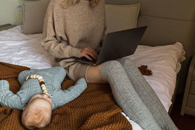 Midsection of mother using laptop while sitting beside sleeping baby on bed at home