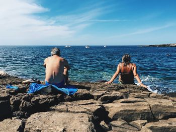 Rear view of men sitting on rock by sea against sky
