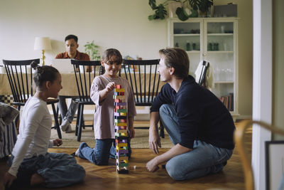 Smiling girl stacking jenga blocks while kneeling on floor by father and sister at home