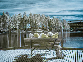 Empty chairs and tables by lake against sky during winter