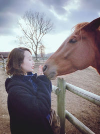 Side view of young woman standing by horse in ranch against sky