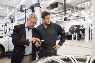 Customer using mobile phone while standing with mechanic in auto repair shop