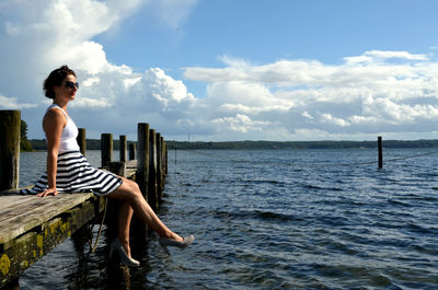 Side view of woman sitting on pier in sea against cloudy sky