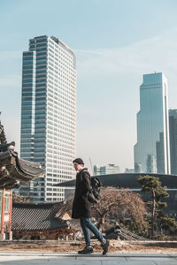 Young man in a hat, black coat and jeans walks on the street. skyscrapers and asian temple 