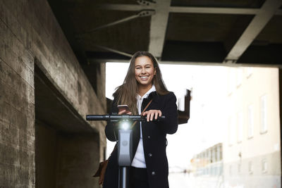 Portrait of smiling teenage girl with electric push scooter holding mobile phone below bridge