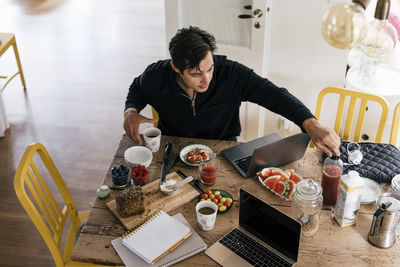 High angle view of male freelancer having breakfast while sitting with laptop on dining table at home