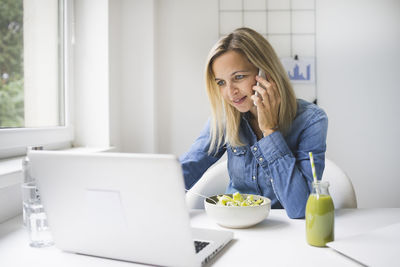 Businesswoman talking on phone while working on laptop at home