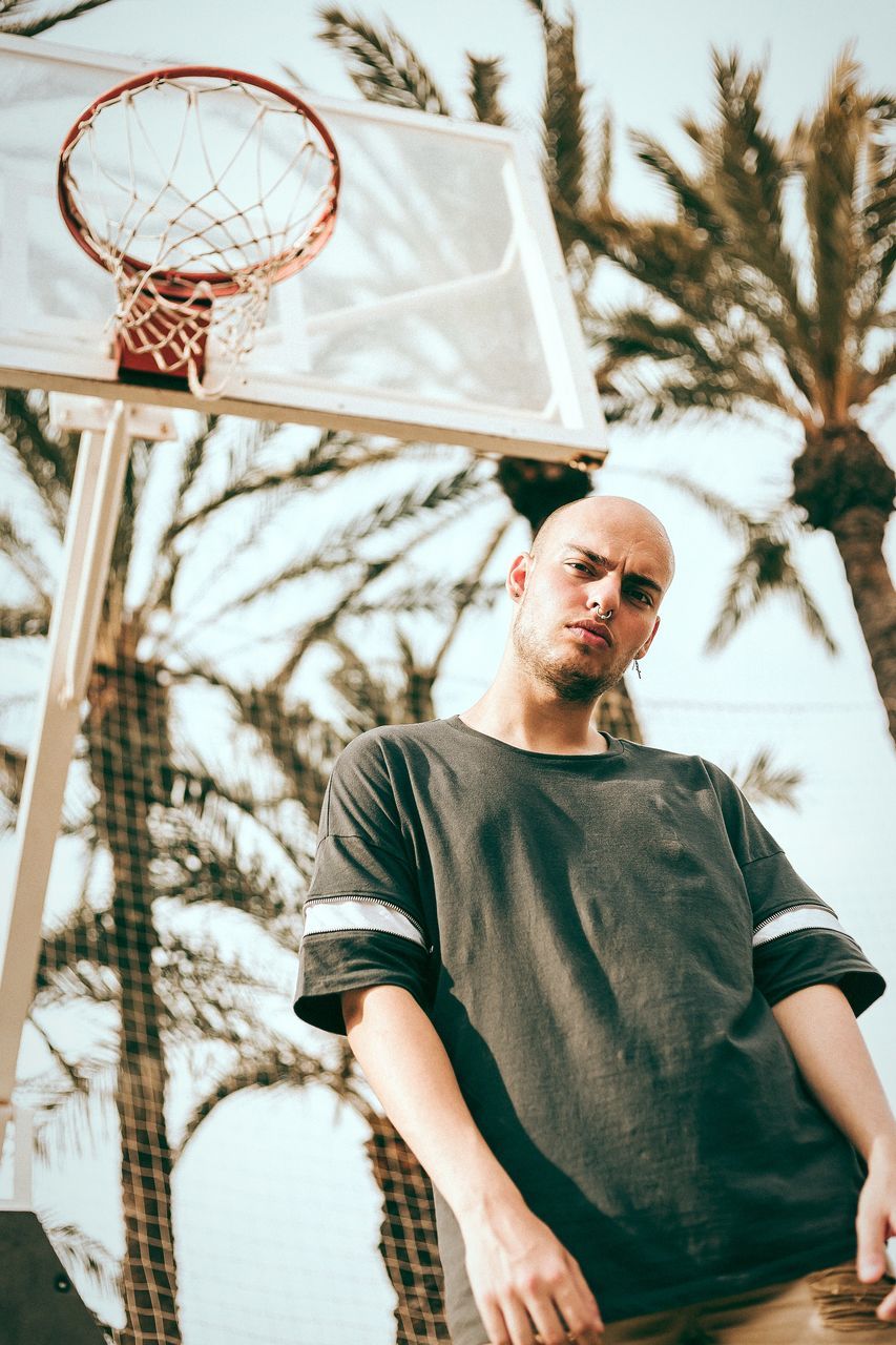 basketball - sport, basketball hoop, basketball player, leisure activity, one person, sport, lifestyles, real people, young men, low angle view, men, court, young adult, standing, outdoors, playing, day, tree