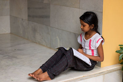Full length of a girl sitting on floor against wall by reading book