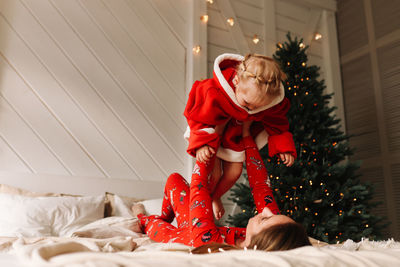 Funny mom and daughter kid in red pajamas having fun on the bed in the room with a christmas tree