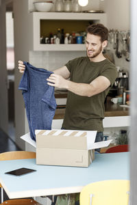 Smiling young man at home unpacking parcel with clothing