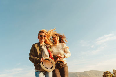 Low angle of laughing women walking together on green meadow in mountain valley in sunlight